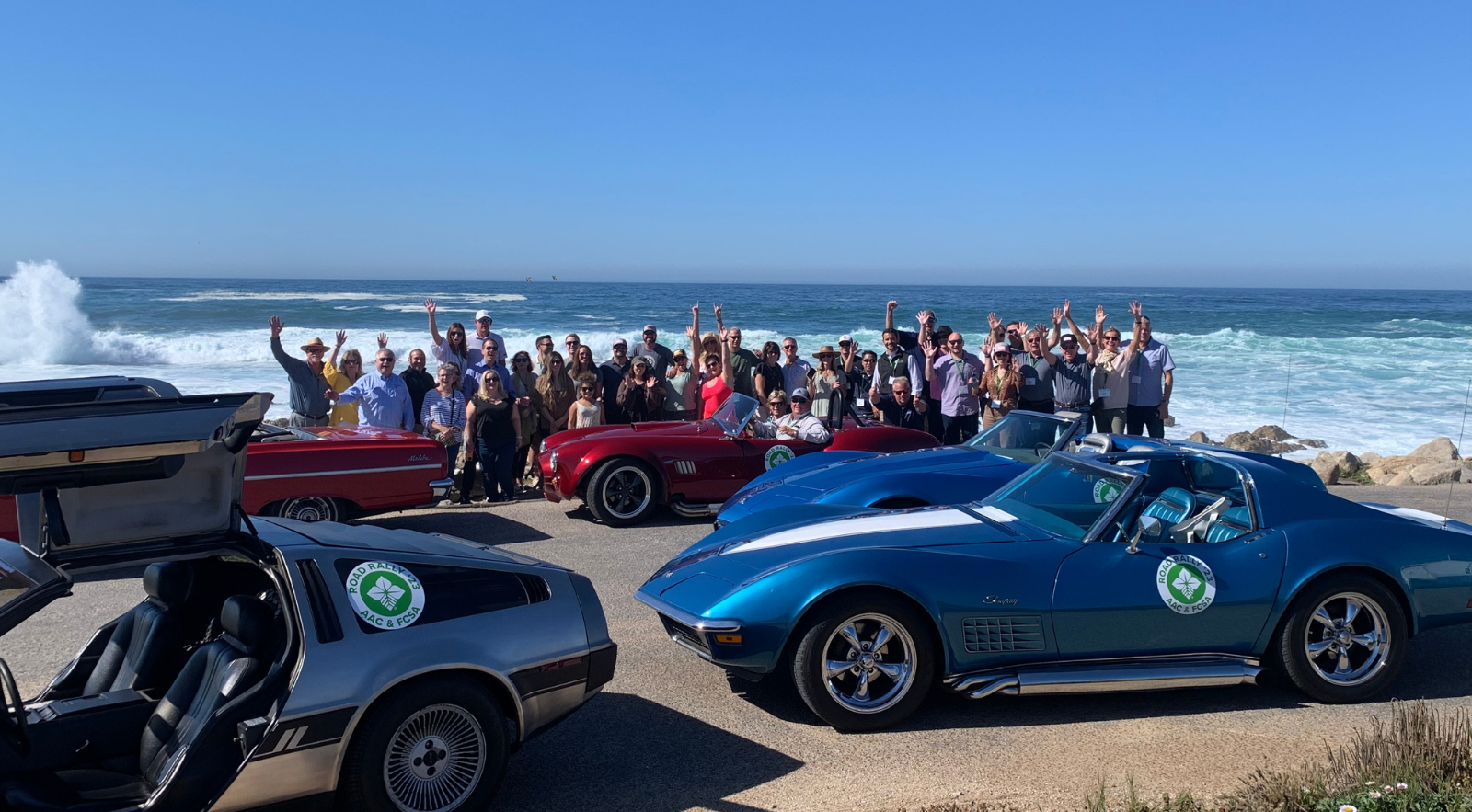 a corporate group standing with classic cars with the pacific ocean in the background on a team-building activity in monterey